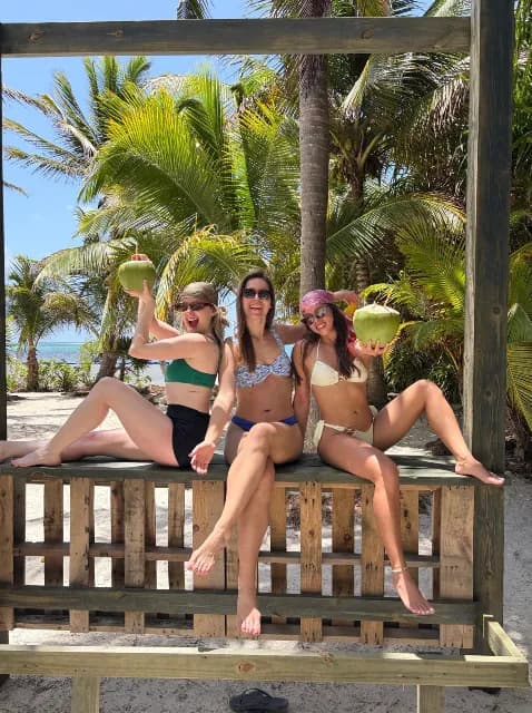 Three friends on a wooden bench under the palms, holding coconuts