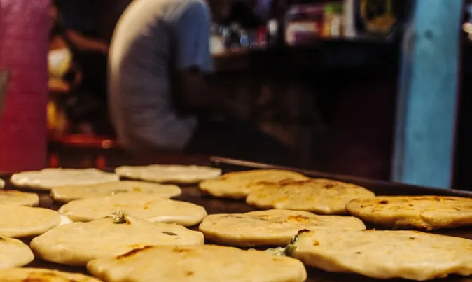 Rounds of corn dough browning on a street griddle, with a cook working behind them