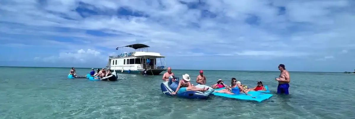 A two-deck houseboat moored in shallow turquoise water, with guests floating beside it