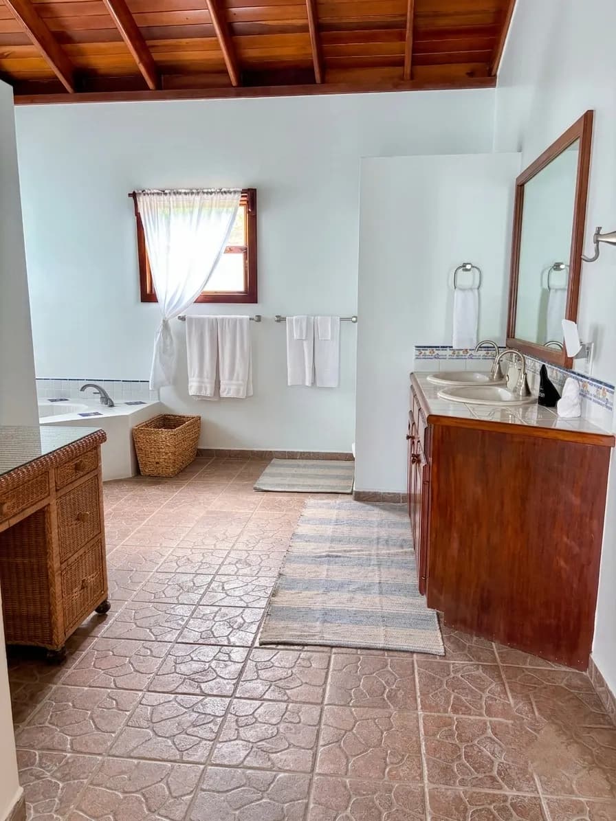 A tiled bathroom under a wood-beamed ceiling, with a corner tub, a wood vanity and towels on a rail
