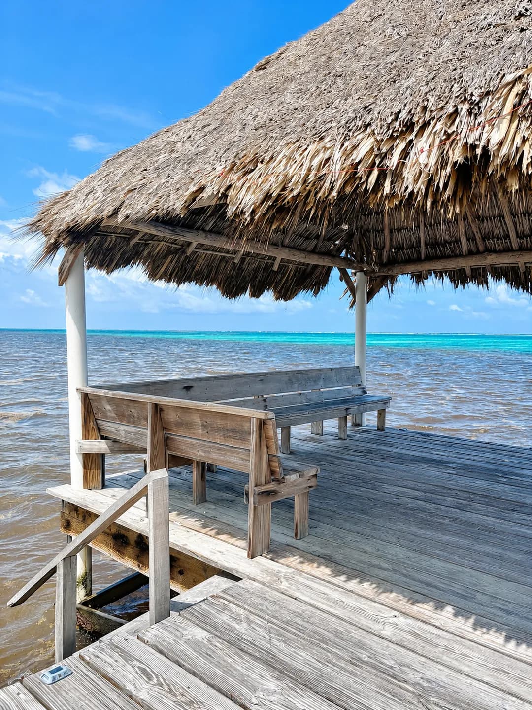 A wooden bench under the thatched palapa at the end of the pier, facing the sea